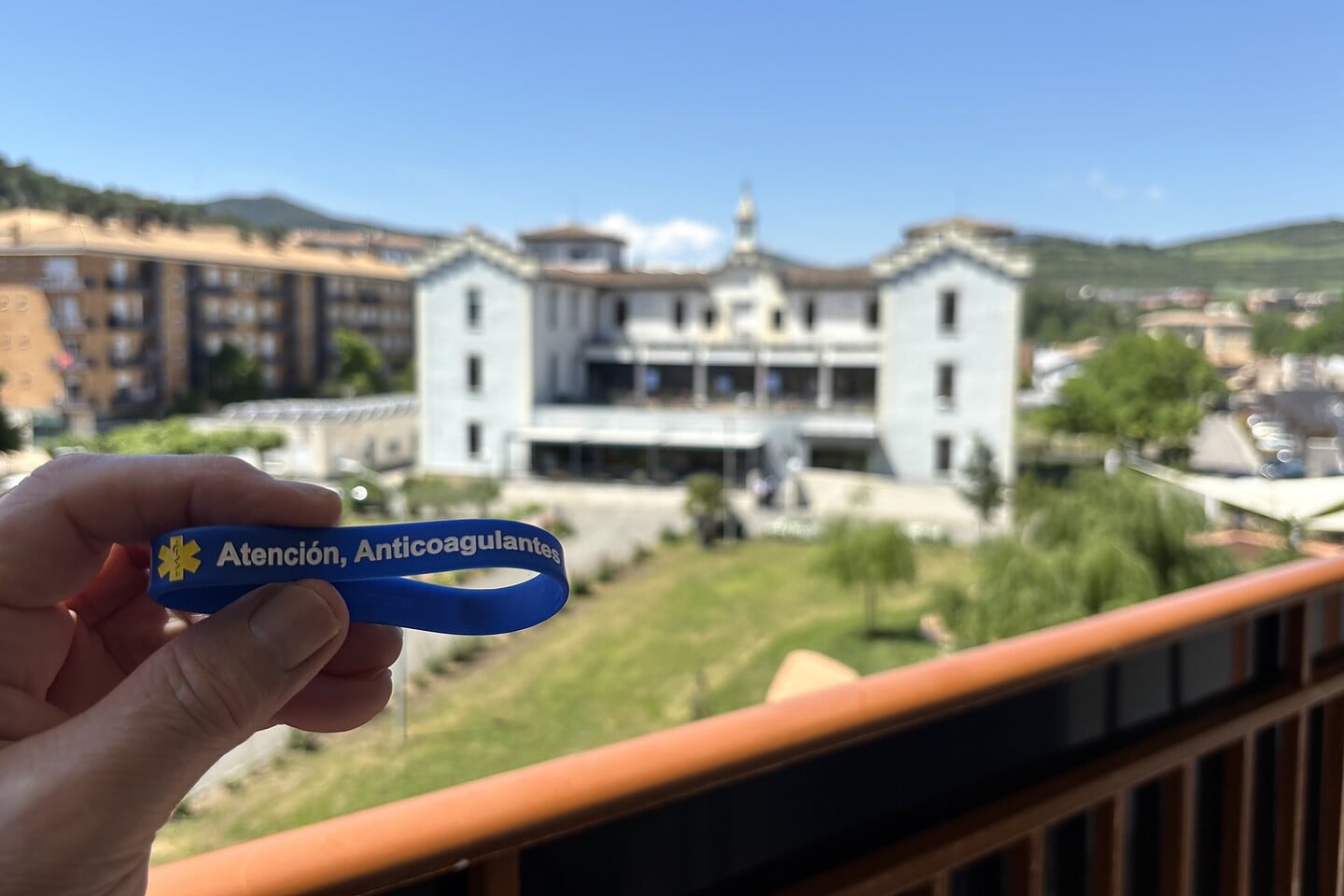 Hand holding a blue Mediband silicone bracelet engraved 'Atención, Anticoagulantes' against a Spanish town backdrop — a traveller's medical ID in the local language